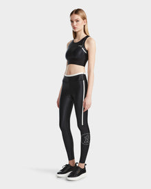 A woman stands against a plain background wearing the Rudsak TRACY - 8524016 Black racer-back sports bra and matching high-waisted leggings with white accents, paired with black sneakers.