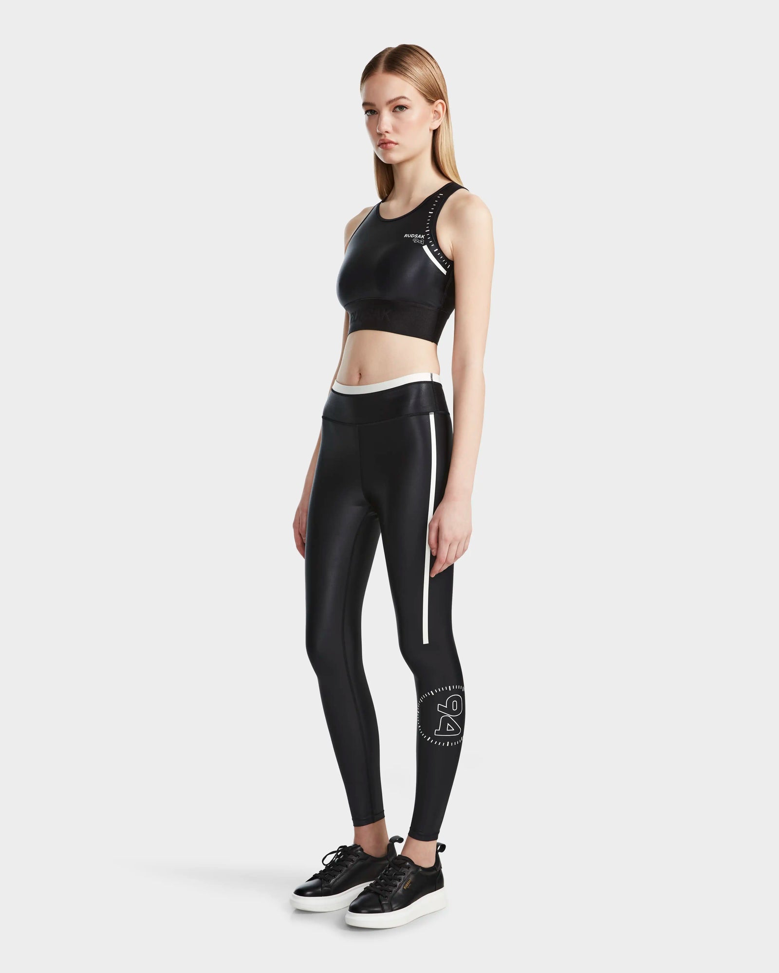 A woman stands against a plain background wearing the Rudsak TRACY - 8524016 Black racer-back sports bra and matching high-waisted leggings with white accents, paired with black sneakers.