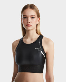 A young woman with straight, blonde hair wears the Rudsak TRACY - 8524016 Black high-performance racer-back sports bra with white logo details, standing against a plain white background and looking at the camera.