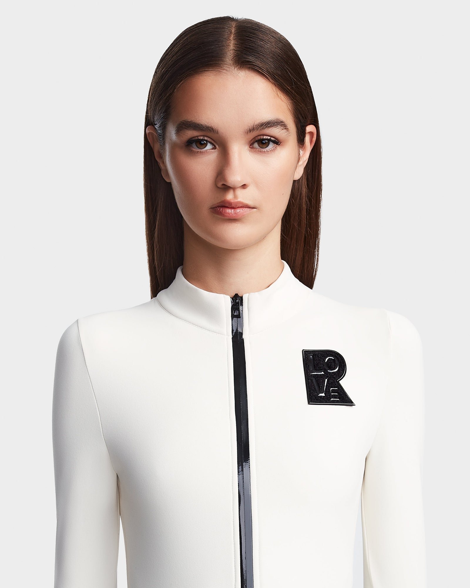 A woman with straight brown hair wears the sleek white Rudsak DAVINA - 8523534 Iceland apres-ski outfit, featuring a black zipper and LOVE brooch, standing against a plain light background and looking directly at the camera.