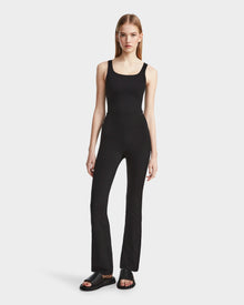 A woman with straight blonde hair and a neutral expression wears Rudsaks ADRIANA - 8524009 Black fitted technical flared-leg leggings, a black tank top, and black slide sandals against a plain white background.