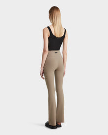 A woman with straight blonde hair is seen from behind wearing the Rudsak ADRIANA - 8524009 Cement taupe flared-leg leggings, a black sleeveless top, and black shoes, standing against a plain white background.