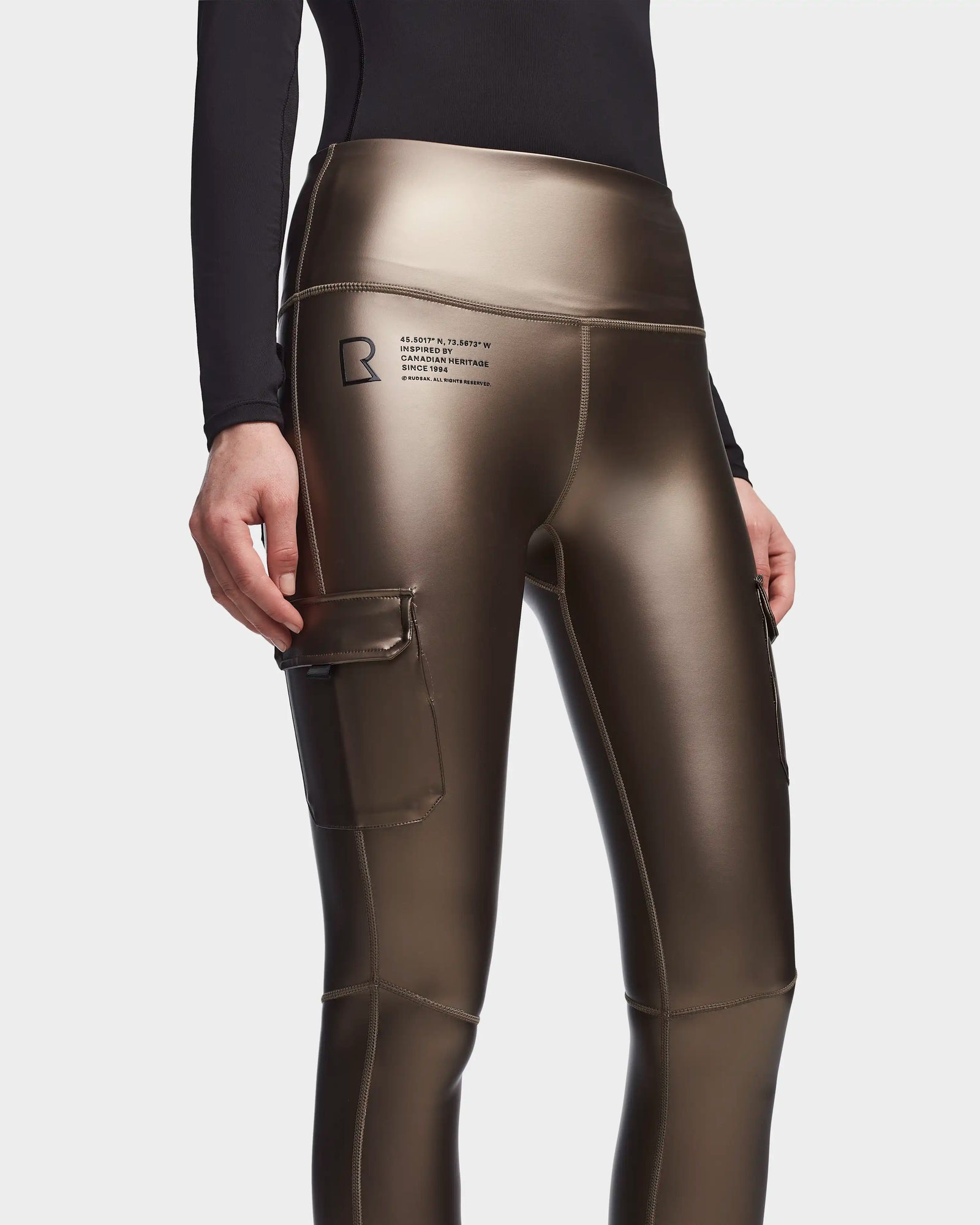Wearing Rudsak SIEN P - 8523027 METALLIC CHOCOLATE stretch vegan leather cargo leggings with a side pocket, Rudsak text and logo on the upper thigh, and a black long-sleeve top.