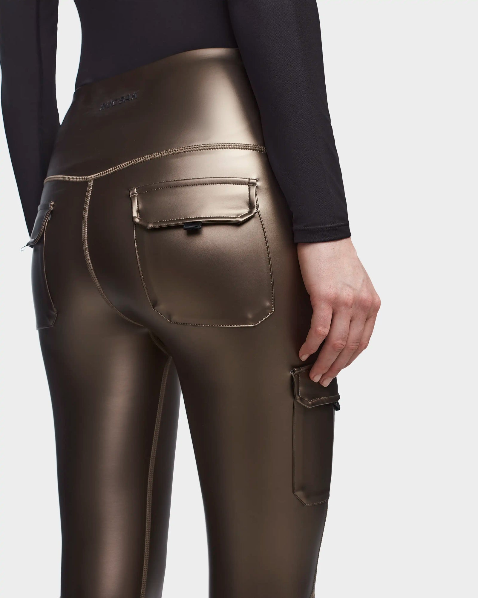 A person is shown from the waist down, standing against a plain light background, wearing Rudsak SIEN P - 8523027 METALLIC CHOCOLATE stretch vegan leather cargo leggings with shiny metallic finish and flap pockets at the back and side.