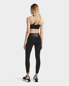 A woman with straight, light brown hair wears Rudsaks THEA - 8523003 BLACK high-rise leggings with a vegan leather waistband and a black sports bra, facing away from the camera against a plain white background.