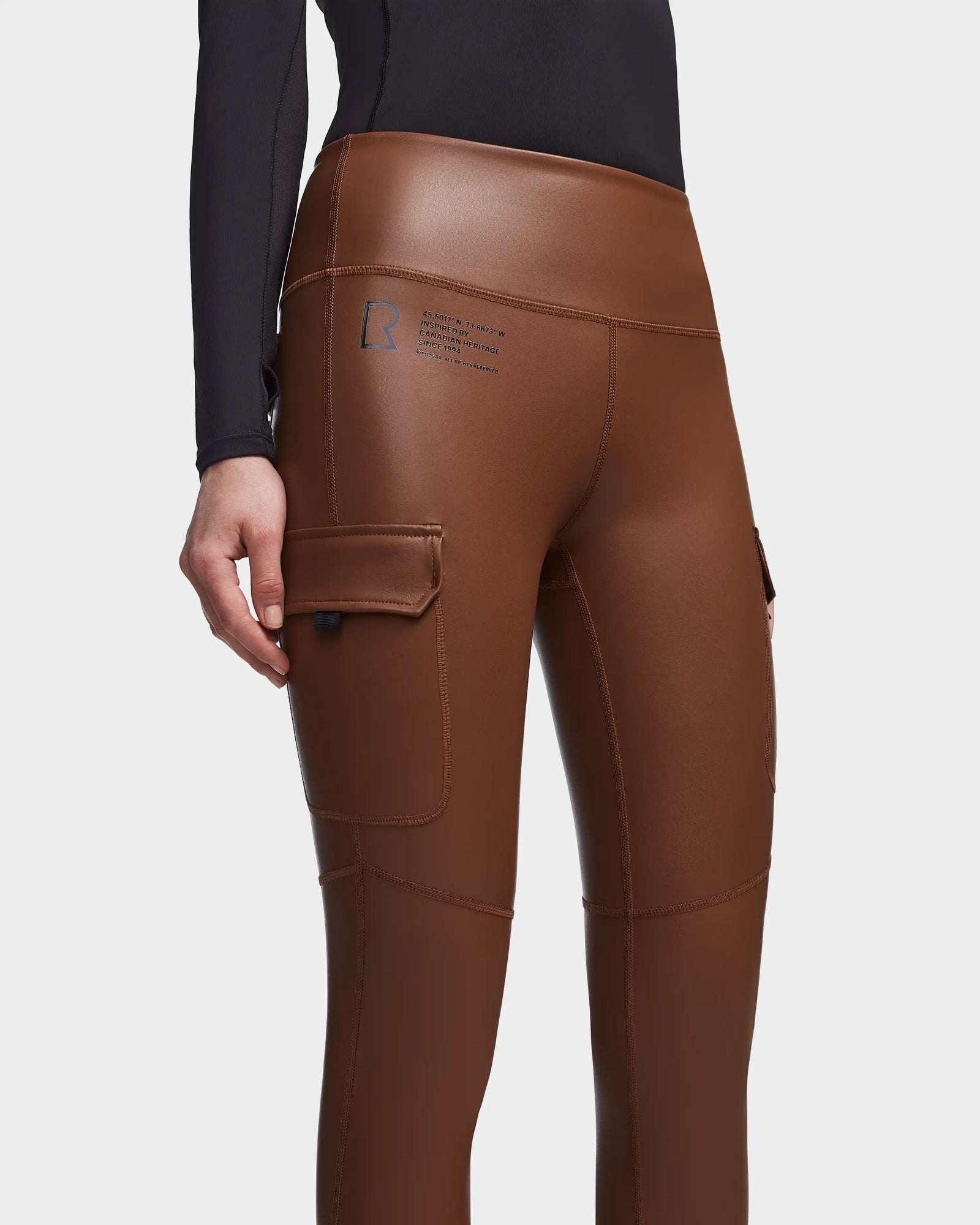 A person wears Rudsak's SIEN L - 8521042 COHIBA shiny brown vegan leather leggings, featuring a side cargo pocket and small printed logo on the upper thigh, paired with a long-sleeve black top—perfect activewear essentials. Only waist to mid-thighs visible.