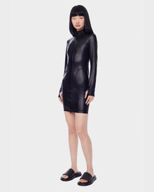 A woman with straight black hair wears the SHANA - 8522508 BLACK fitted zip-up stretch vegan leather dress by Rudsak and black slides, standing against a plain white background.