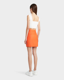 A woman with straight brown hair faces away, wearing a white crop top, white sneakers, and the Rudsak SABINE - 8524063 Orange Sorbet quilted short skirt for a modern look against a plain light background.