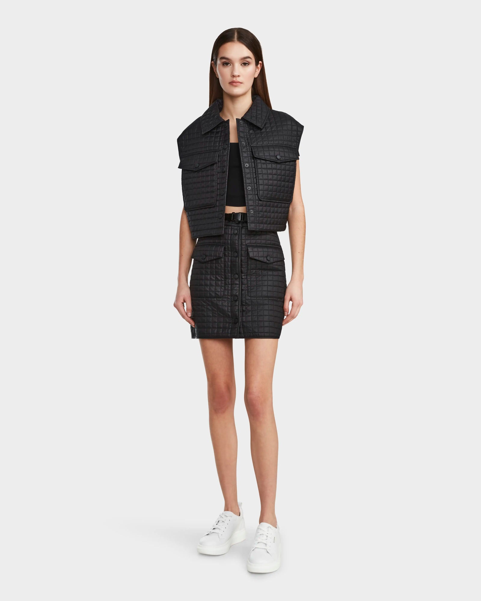 A woman faces forward in a Rudsak SOLSA - 8124048 Black cropped quilted vest with eco-friendly Sorona fill, styled with a matching skirt, black crop top, and white sneakers against a plain white background.