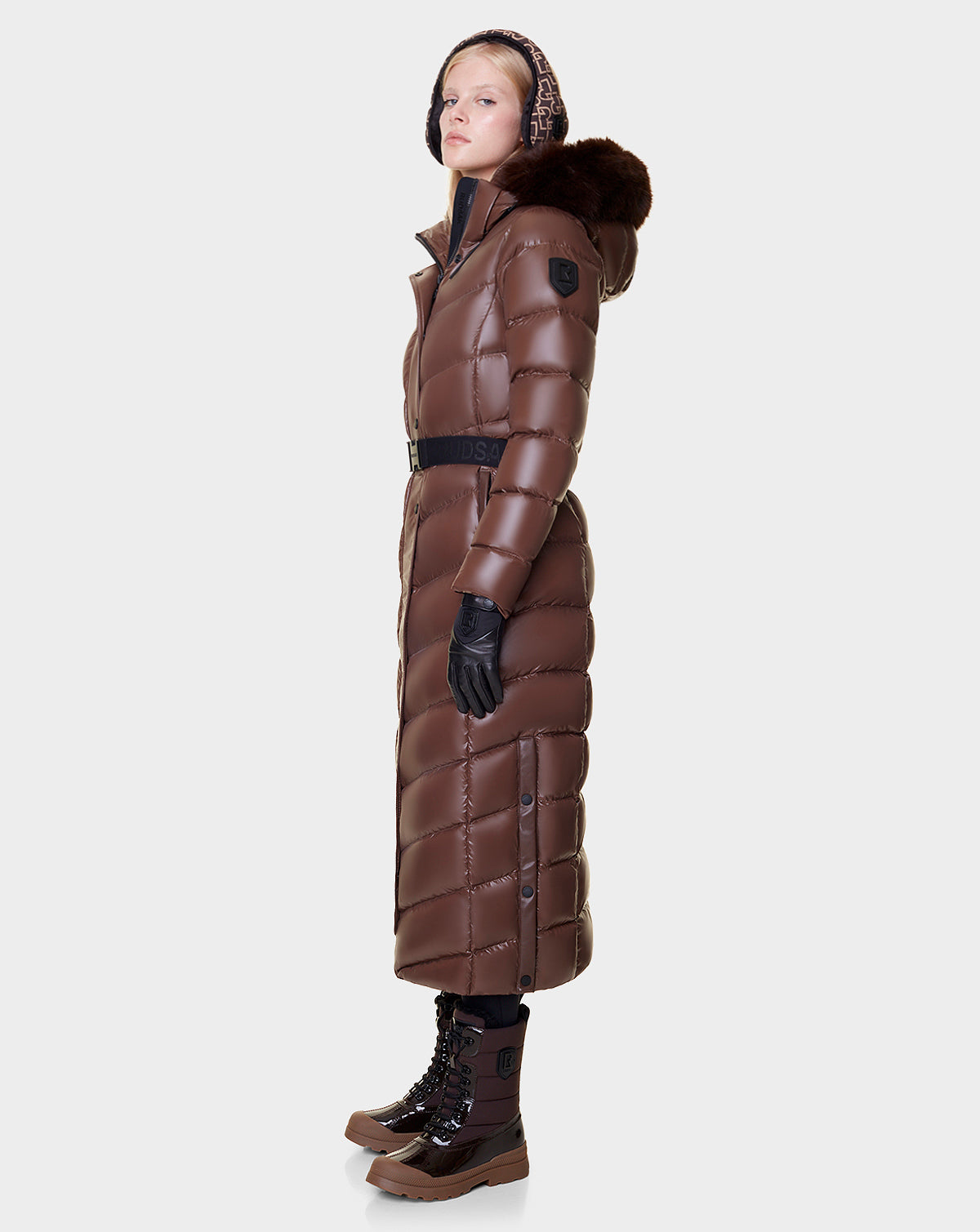 A person in profile wears the Rudsak ELSIE - L121525 CAOCAO, a long brown maxi puffer coat with a fur-trimmed hood, paired with black gloves and brown lace-up winter boots against a plain light backdrop.