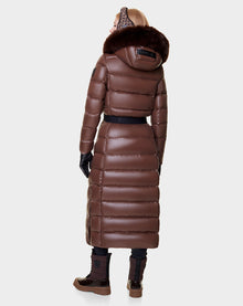 A person in the Rudsak ELSIE - L121525 CAOCAO womens maxi down puffer with RDS-certified down, a shiny brown coat with a removable fur-trimmed hood, black gloves, and brown boots stands facing away against a plain light background.
