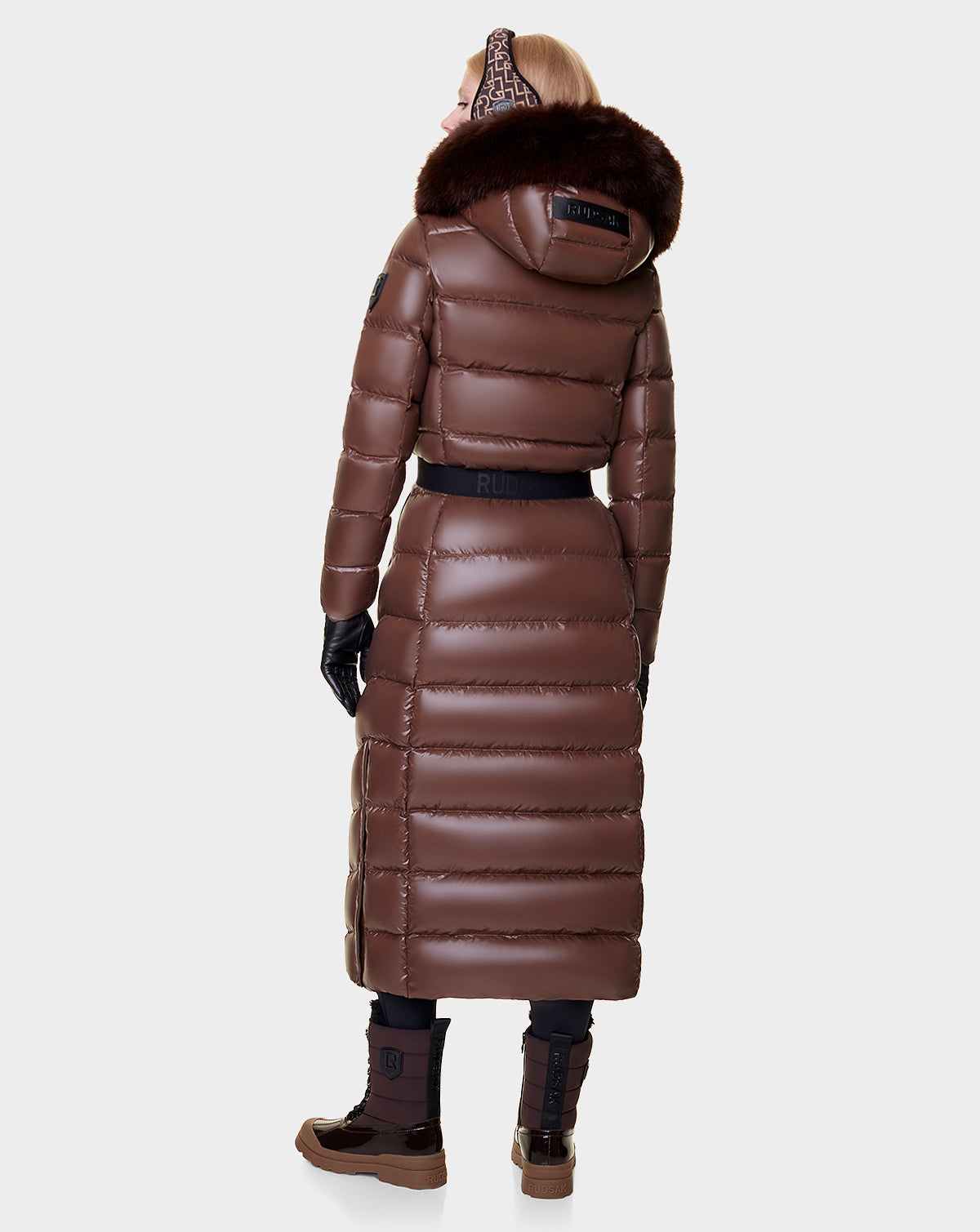 A person in the Rudsak ELSIE - L121525 CAOCAO womens maxi down puffer with RDS-certified down, a shiny brown coat with a removable fur-trimmed hood, black gloves, and brown boots stands facing away against a plain light background.