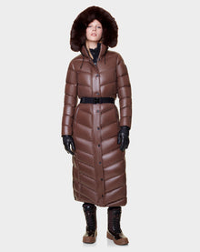 A person stands against a plain light background wearing the Rudsak ELSIE - L121525 CAOCAO womens maxi down puffer with a fur-lined removable hood, black belt, black gloves, and brown winter boots.