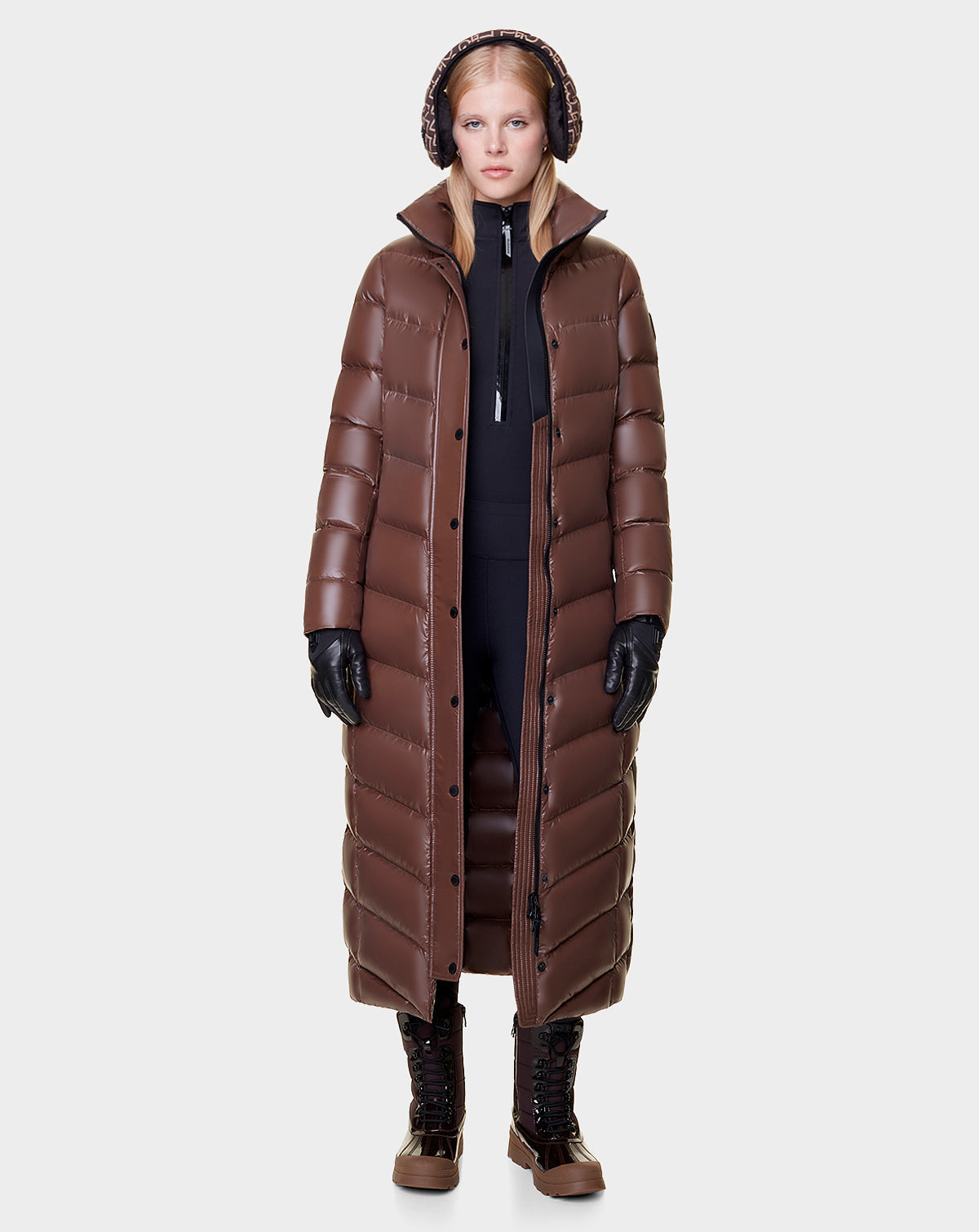 A woman faces forward in the Rudsak ELSIE - L121525 CAOCAO womens maxi down puffer with RDS-certified down, showcasing a long brown silhouette, high collar, black gloves, leggings, brown boots, and earmuffs against a light background.