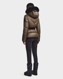 A person stands facing away, wearing the Rudsak CHRISTELLE S - L123524 Light Olive puffer jacket with a black fur-trimmed hood, paired with black leggings and winter boots against a plain light background.