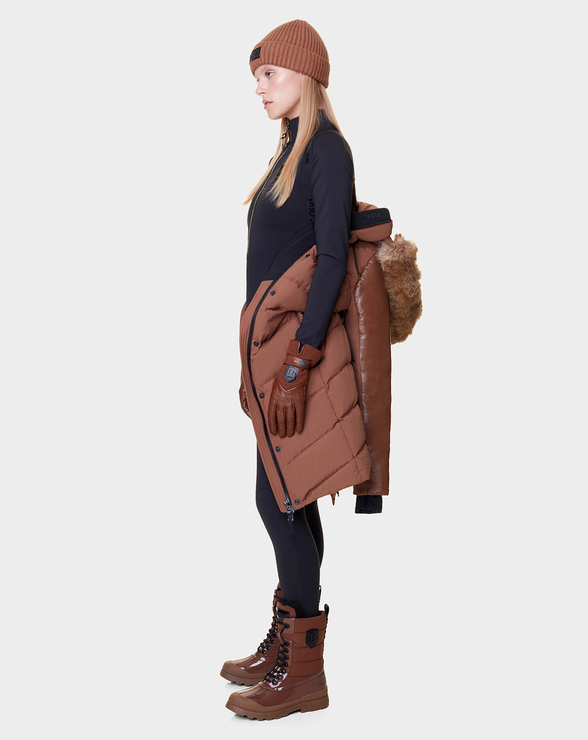 A woman in black, brown winter boots, beanie, and gloves stands in profile holding the Rudsak ELSA - L121519 TAUPE women’s quilted down puffer with fur hood—crafted sustainably—over her shoulder against a plain light background.