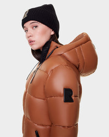 A woman wears the Rudsak EVIE - L122517 DARK CAMEL down puffer with a hood and waterproof zippers, both showcasing the bold R logo, as she looks over her shoulder against a plain light background.