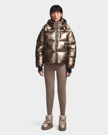A person wearing the Rudsak MAVE S - 8123563 Metallic Ivory Camo womens down puffer jacket, taupe leggings, and matching gold winter boots stands against a plain white background.