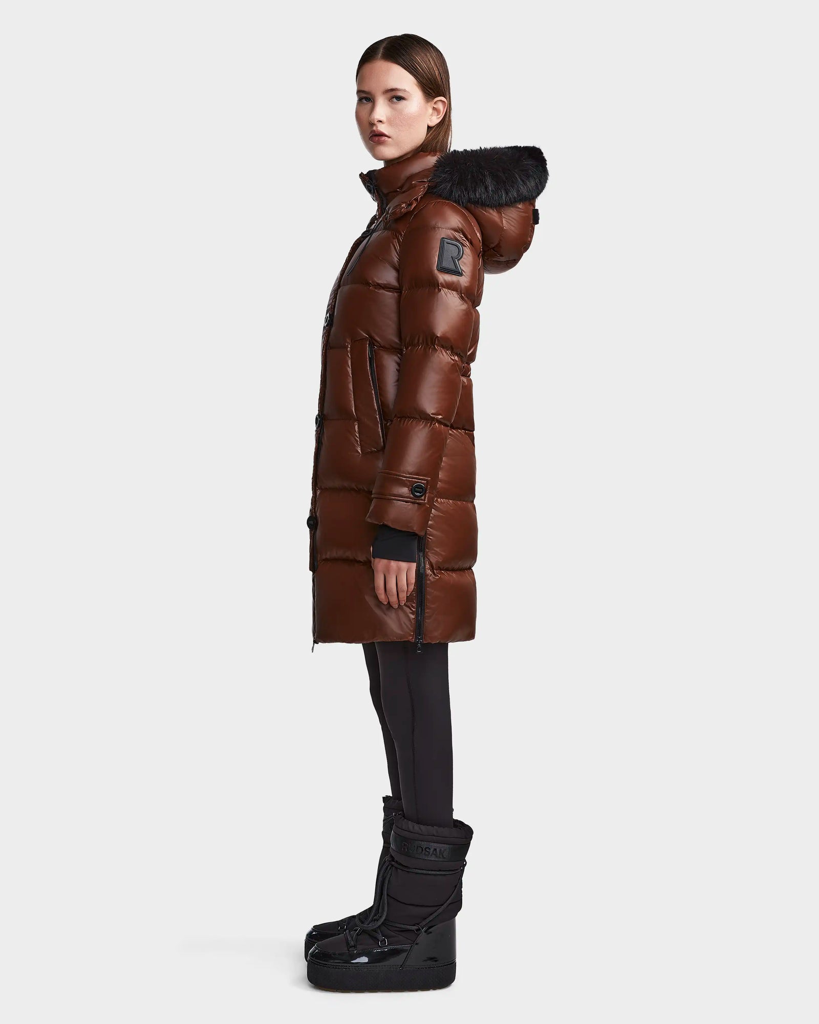 A woman stands in profile wearing the Rudsak MEYA S - L123521 Cohiba, a long brown women’s down puffer with a black fur-trimmed removable hood, paired with black leggings and winter boots against a plain white background.