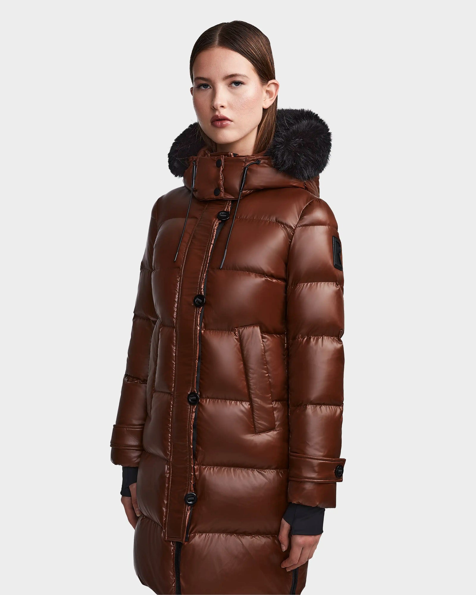 A woman models the Rudsak MEYA S - L123521 Cohiba, a shiny brown down puffer coat with a high collar, black fur-trimmed removable hood, and button details, standing against a plain light gray background.