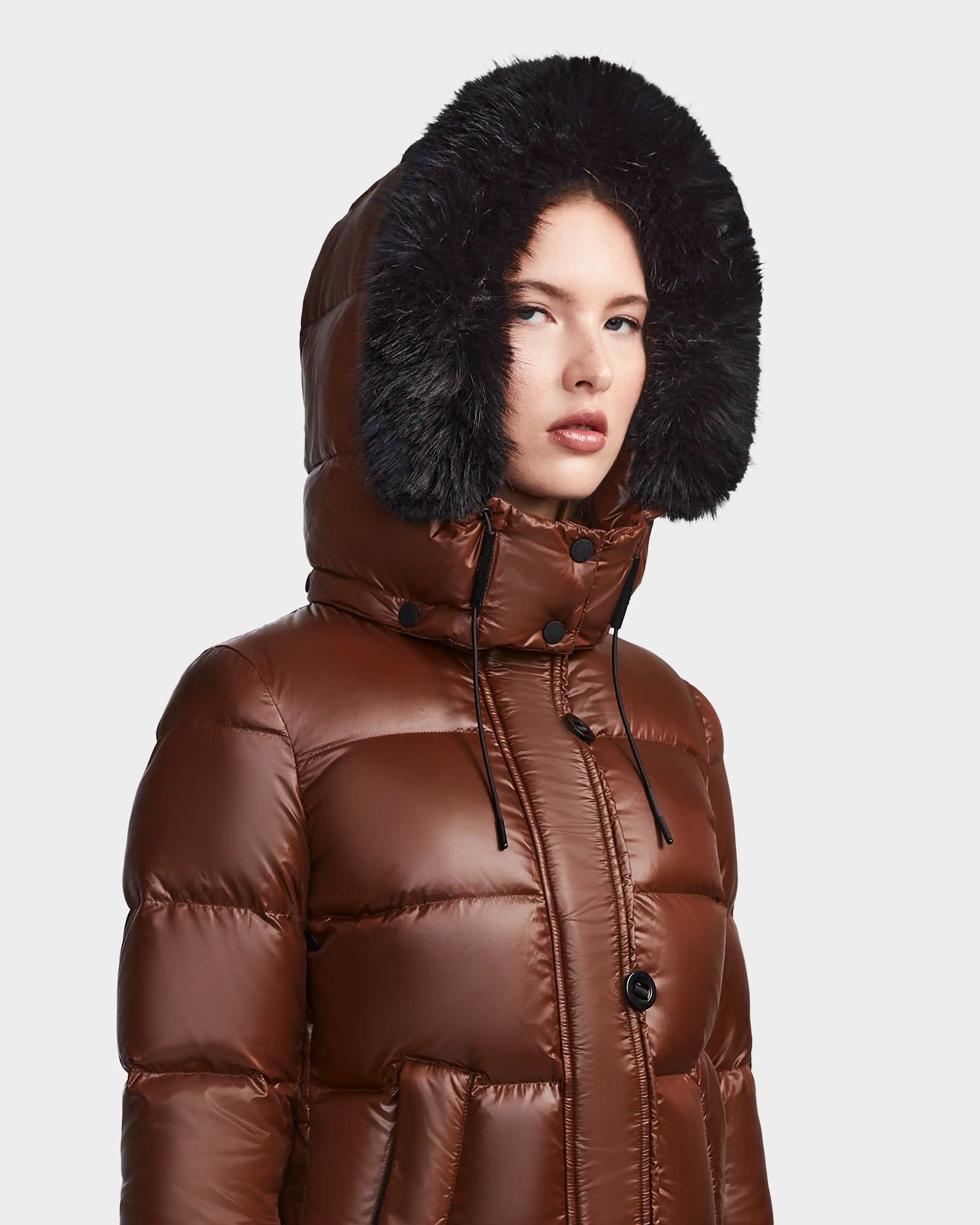 A person wears the Rudsak MEYA S - L123521 Cohiba, a shiny brown women's down puffer jacket with a large black faux fur-trimmed removable hood, looking slightly to the side against a plain light background.