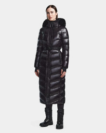 A woman stands against a white background wearing the Rudsak ELSIE S - L123518 Black maxi down puffer with a belted waist and fur-trimmed hood, styled with black leggings and lace-up boots.