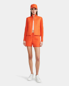 A woman faces forward against a white background, wearing the Rudsak LOUISE - 8124046 jacket in Orange Sorbet, matching shorts and cap, paired with a white crop top and sneakers.