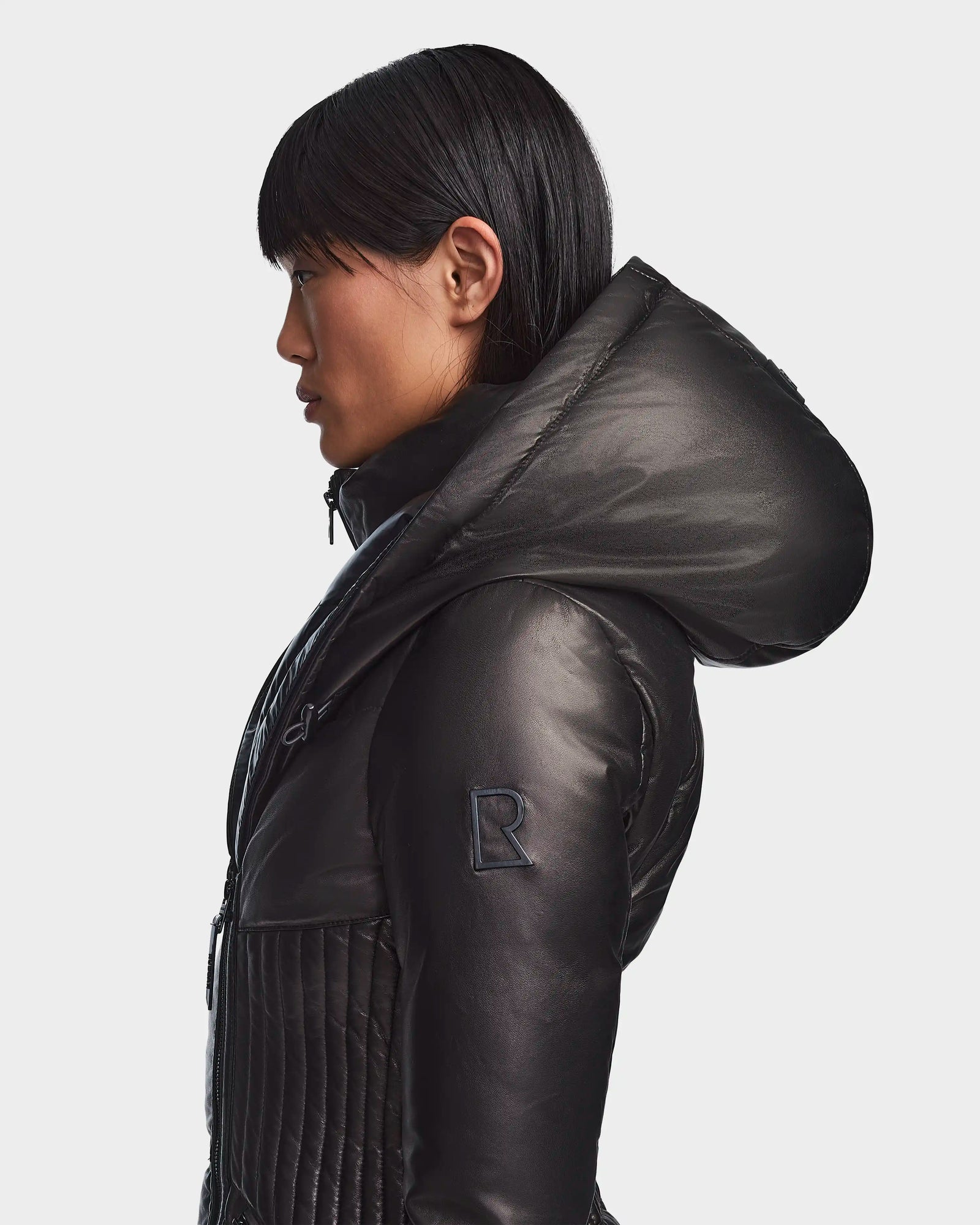 A woman with straight dark hair wears the SHAUNA X - 8121568 BLACK puffer jacket by Rudsak, shown in profile against a light background. The shiny black jacket has a prominent hood and a sleeve patch with the letter R.