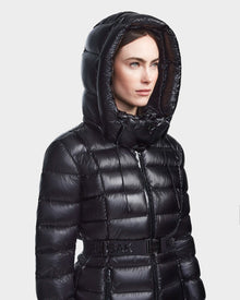 A woman wears the Rudsak FRIDA S - L123513, a shiny black mid-length down jacket with horizontal quilting and a belted waist, hood up, standing against a plain light background.