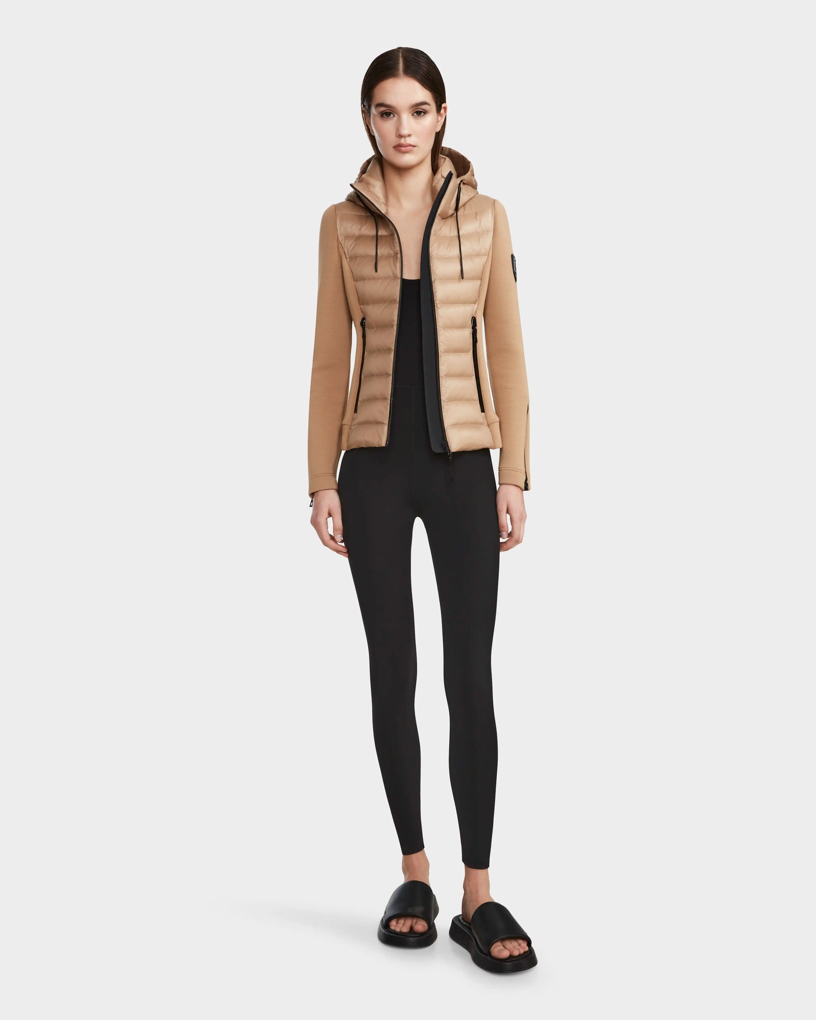 A woman faces forward in a RUDSAK MABS - 8122020 TOFFEE women’s puffer jacket with a hood, paired with black leggings and black slide sandals. She has straight brown hair pulled back, a neutral expression, and stands against a plain white background.