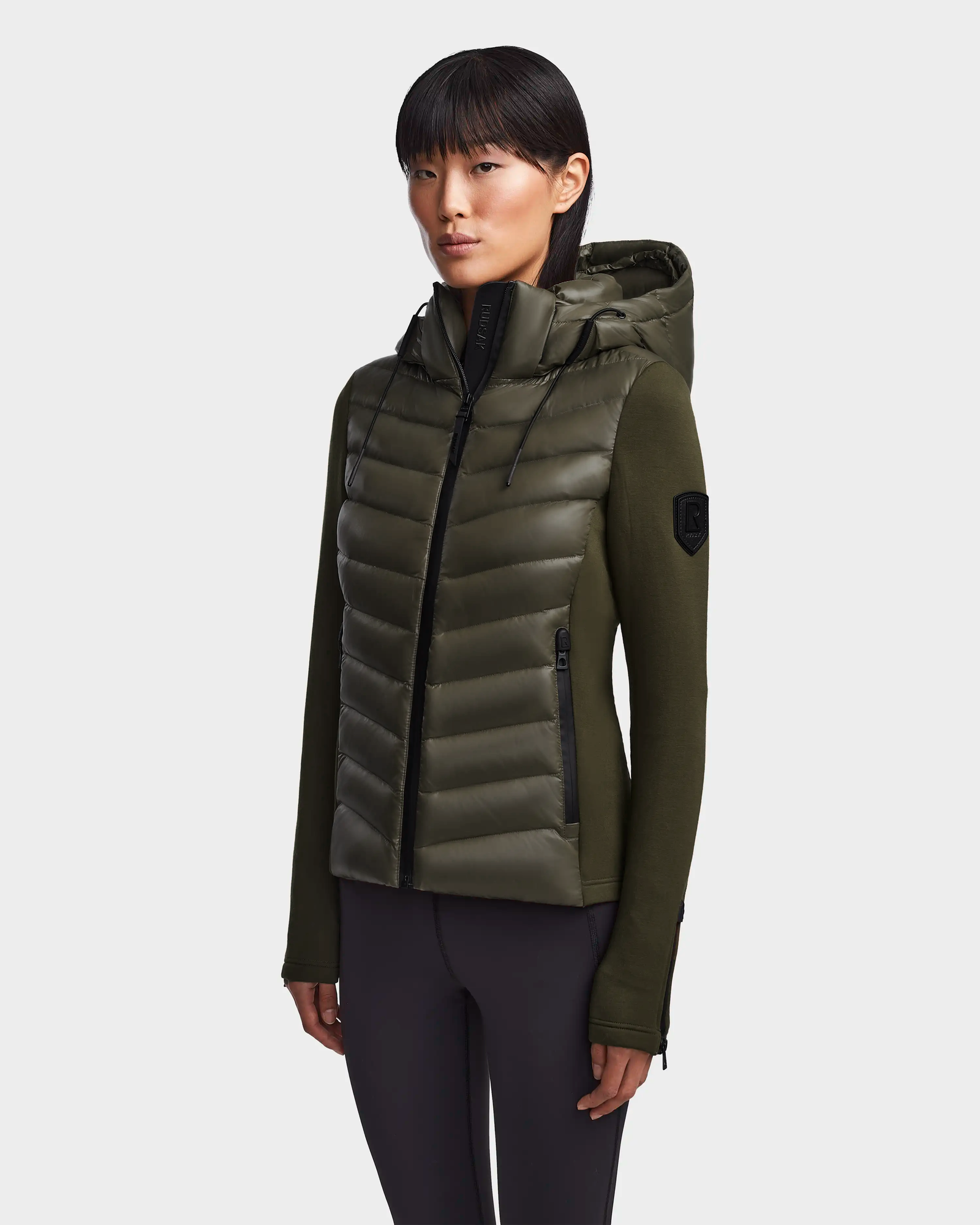 A woman with straight dark hair wears the Rudsak MALYA - L121006 Light Olive down quilted jacket with knit sleeves and black zippers, paired with dark pants, standing against a plain light background.