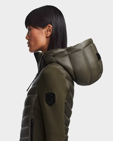 A woman with straight dark hair, seen in profile against a plain light background, wears the Rudsak MALYA - L121006 Light Olive activewear-inspired quilted hooded jacket featuring a black sleeve patch.