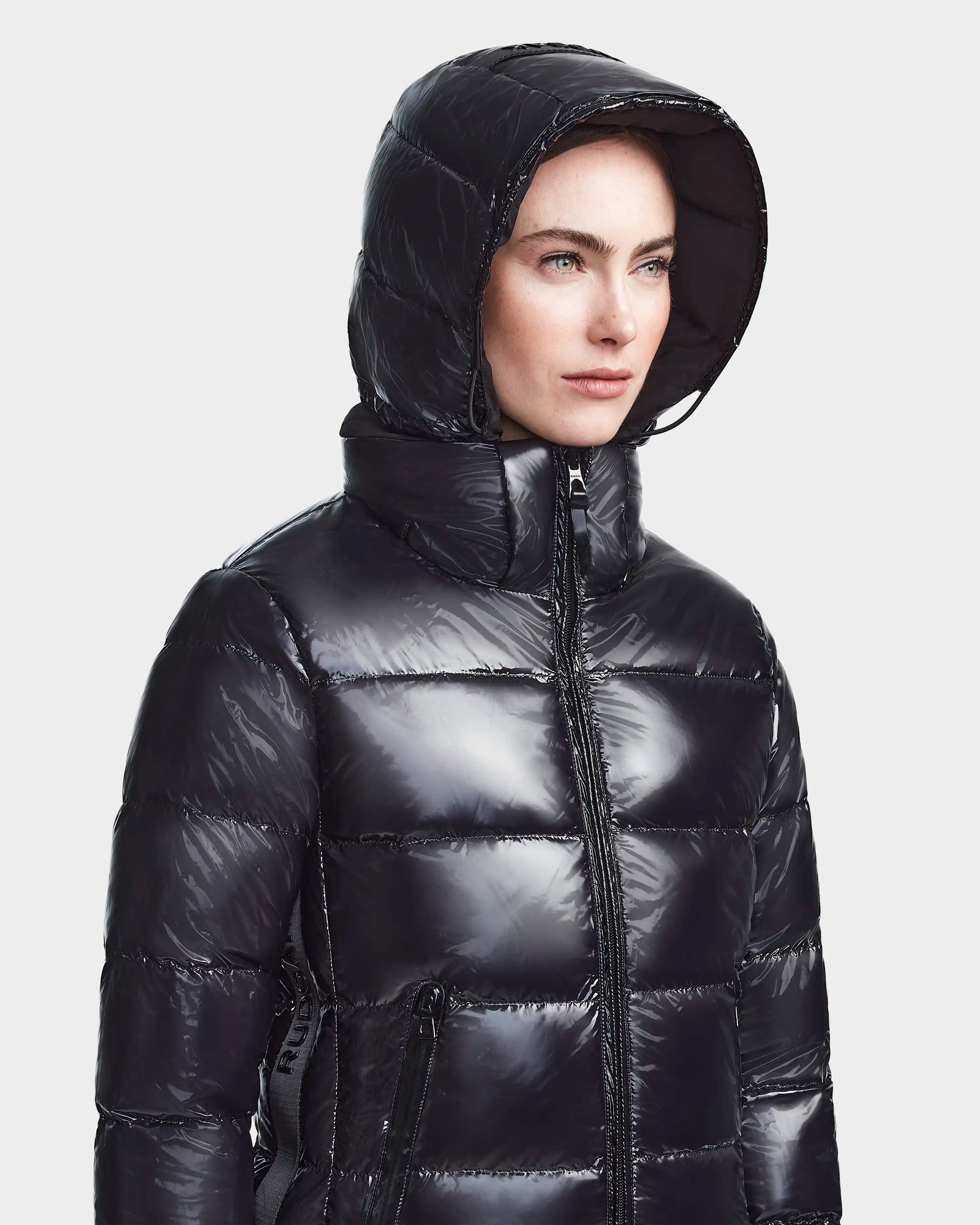 Someone wearing the RUDSAK MARNIE LONG - 8122519 BLACK womens puffer jacket with a hood, facing slightly to the side against a plain light background.