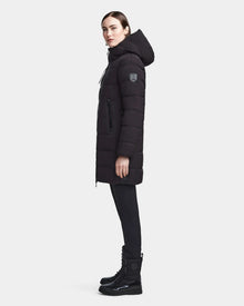 A woman in profile wears the RUDSAK MARNIE LONG - 8122519 BLACK womens puffer jacket with a hood, black leggings, and black lace-up boots against a plain light background.