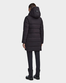 A person is seen from behind, wearing the RUDSAK MARNIE LONG - 8122519 BLACK womens puffer jacket with a hood, paired with black pants and boots, all set against a plain white background.