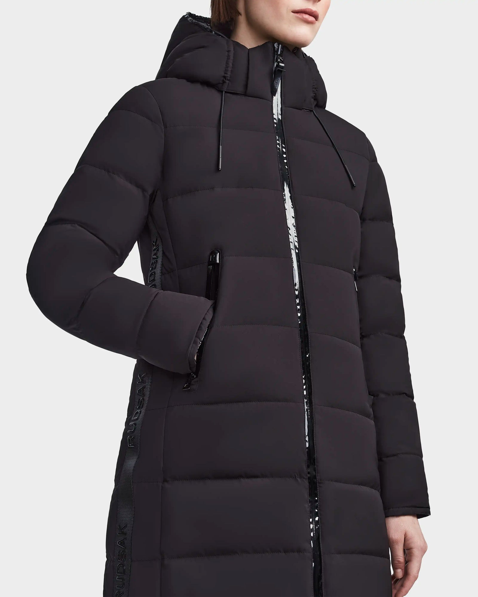 A woman stands against a plain background wearing the RUDSAK MARNIE LONG - 8122519 BLACK jacket, which features a hood, shiny black zipper and trim, and has one hand in a side pocket.