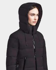 A woman wears the RUDSAK MARNIE LONG - 8122519 BLACK jacket with a hood, front zipper pockets, and drawstrings, looking off to the side against a plain light background.