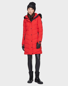 A woman wears the Rudsak ELISA - L121512 POPPY RED sustainable winter jacket—a long red quilted down puffer with fur-trimmed hood—paired with a black knit beanie, gloves, leather leggings, and lace-up boots against a plain light background.