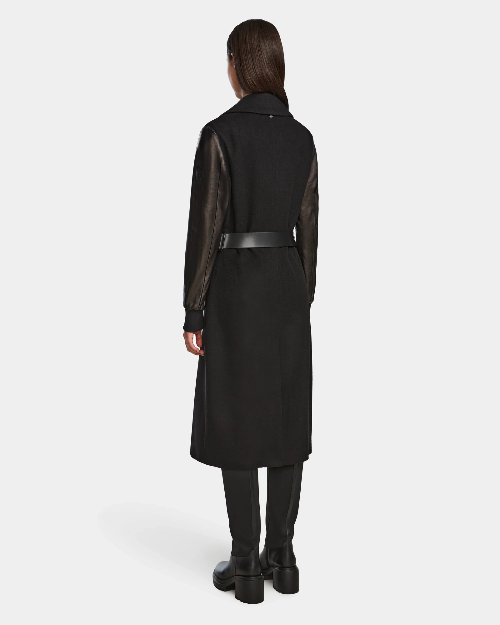 A woman faces away, wearing the Rudsak MARYLIN - 8124520 Black wool coat with leather sleeves, a black belt, black pants, and black boots against a plain white background.