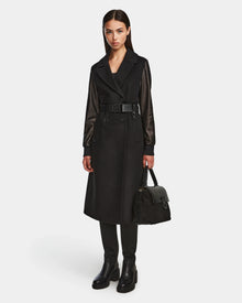 A woman with straight dark hair and a neutral expression stands against a plain background wearing the Rudsak MARYLIN - 8124520 Black wool coat with leather sleeves, paired with black leather boots and holding a black handbag.