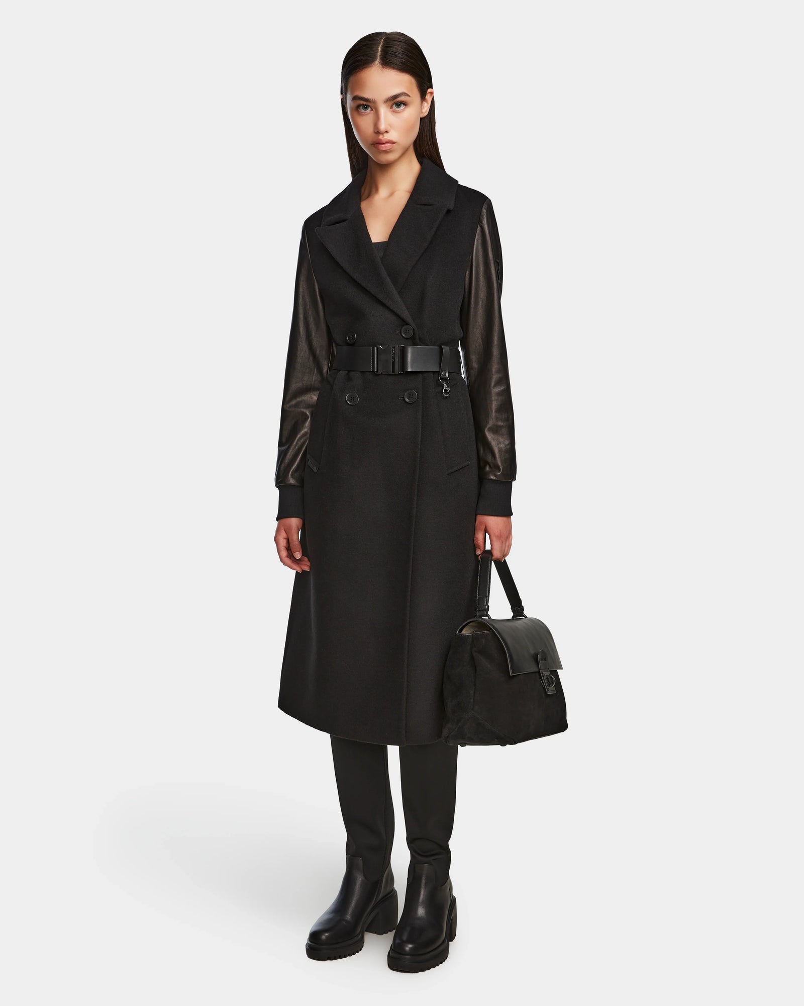 A woman with straight dark hair and a neutral expression stands against a plain background wearing the Rudsak MARYLIN - 8124520 Black wool coat with leather sleeves, paired with black leather boots and holding a black handbag.