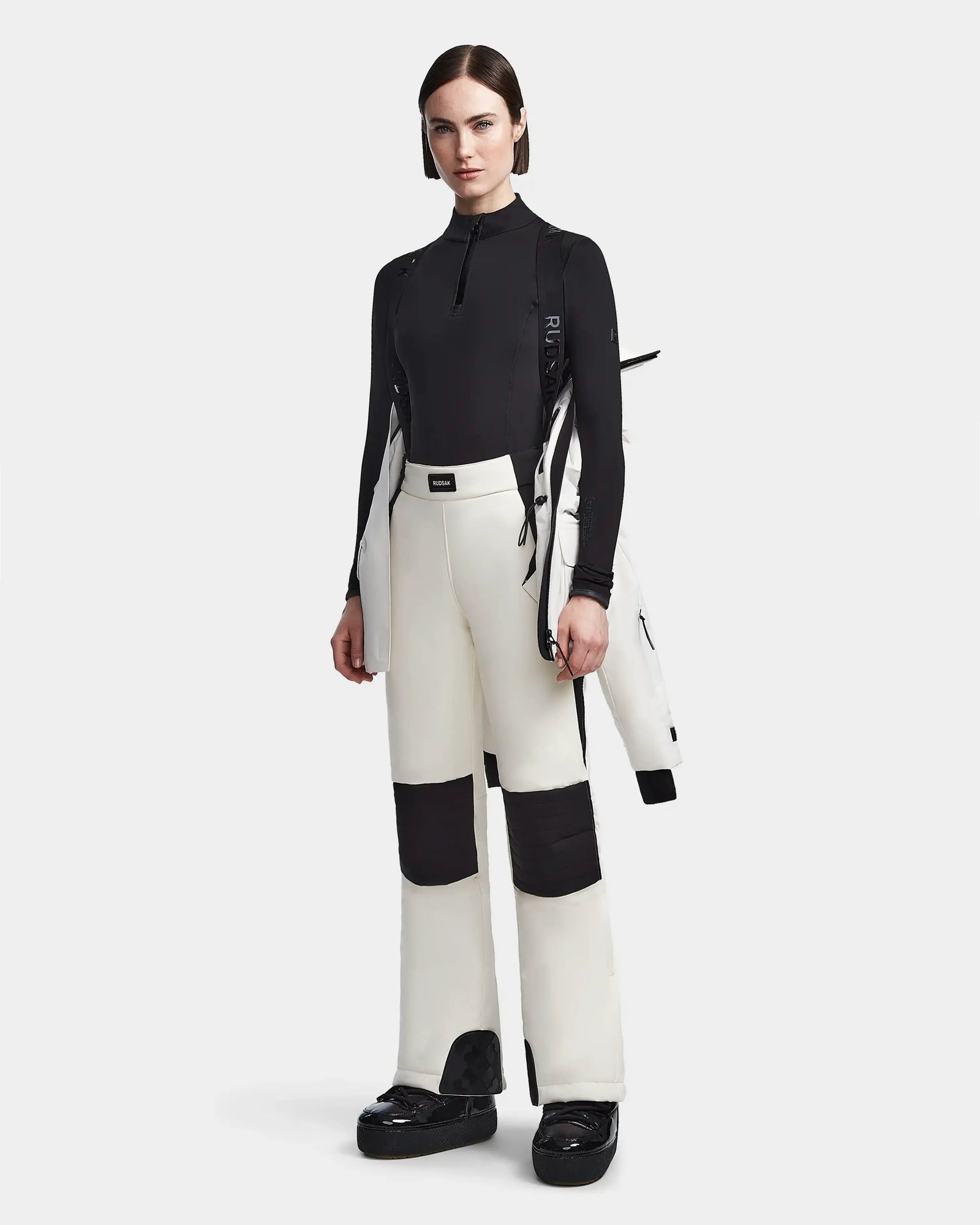 A woman faces forward in a black fitted top, white pants with black knee patches, and black shoes. She holds the Rudsak LUX - 8123618 Iceland waterproof ski jacket over her shoulder, posing against a plain light background.
