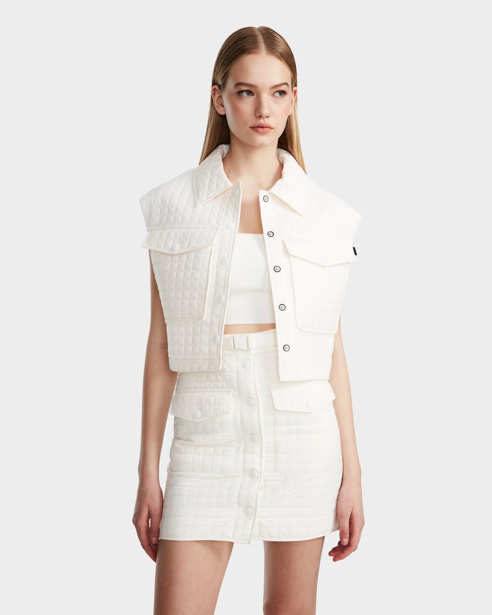 A woman with straight blonde hair wears the Rudsak SOLSA - 8124048 Eggshell womens cropped quilted vest over a white crop top and matching skirt, standing against a light gray background and looking slightly to her left.