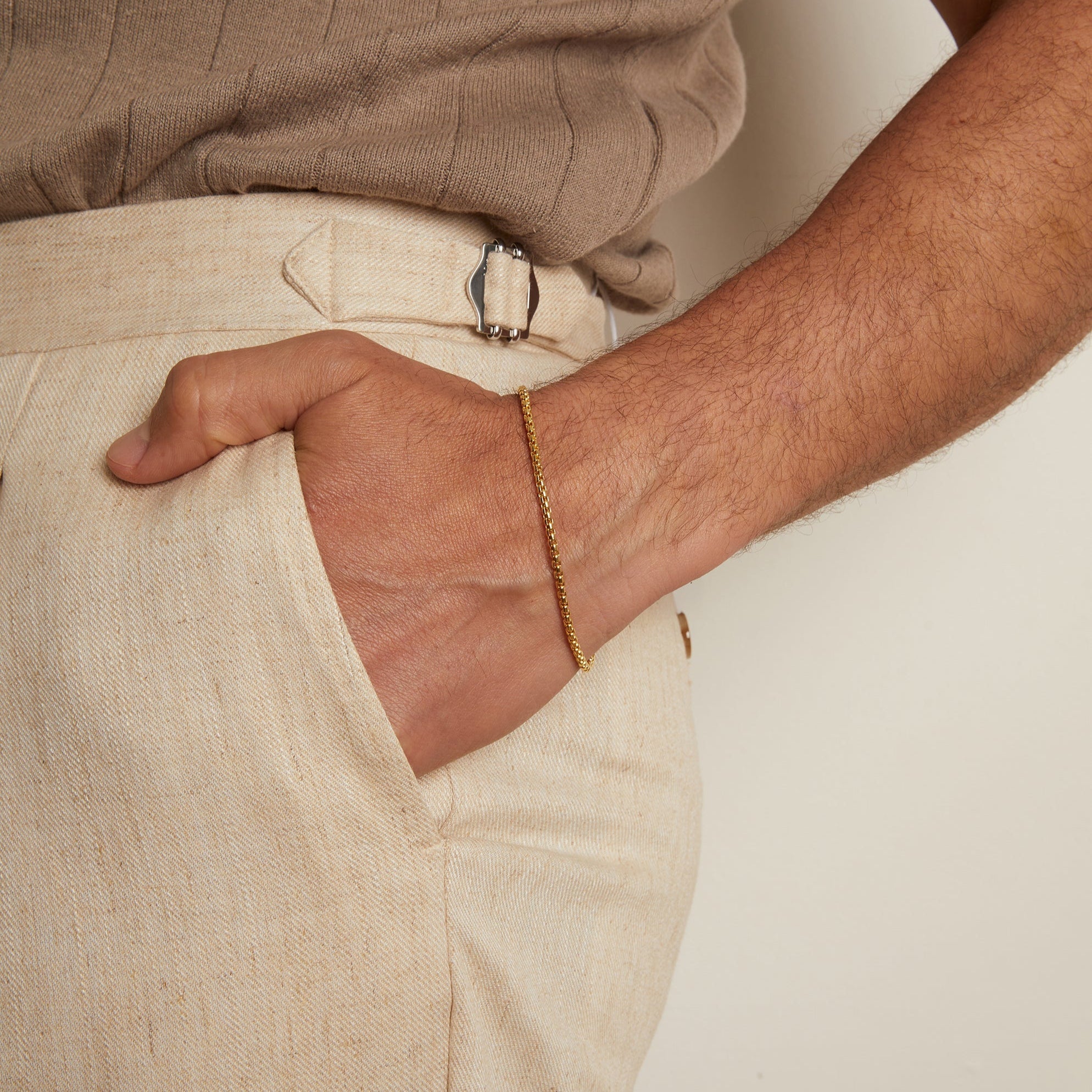 Model. Person wearing beige shirt and pants with a gold bracelet on a plain background