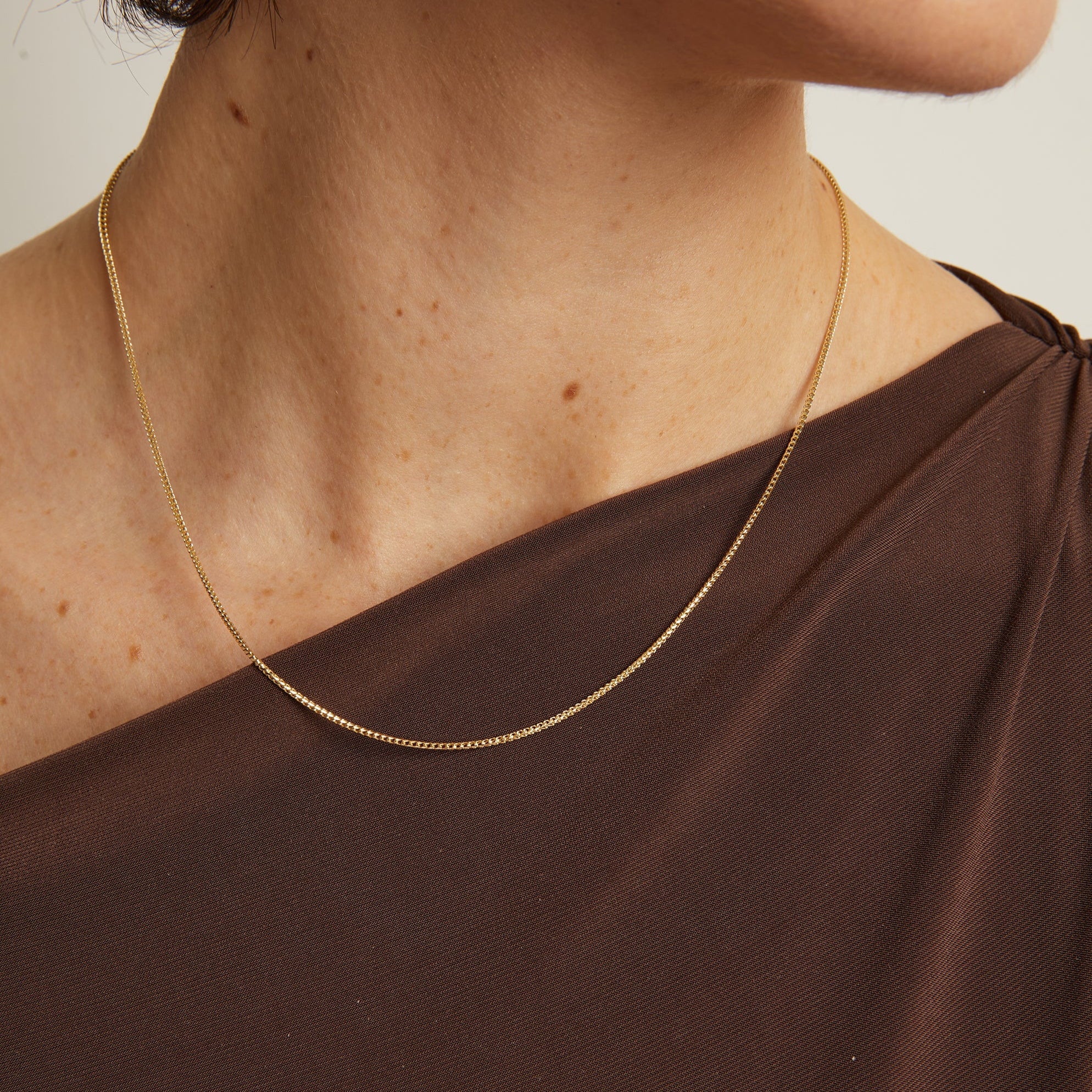 Model. Gold necklace on a person wearing a brown top against a neutral background