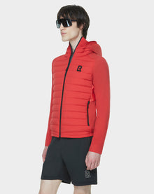 A man wears large black sunglasses, the RUDSAK JARRY - 6122007 POPPY RED RDS-certified down hooded puffer with a black chest logo, and black shorts with a matching R logo, standing against a plain white background.