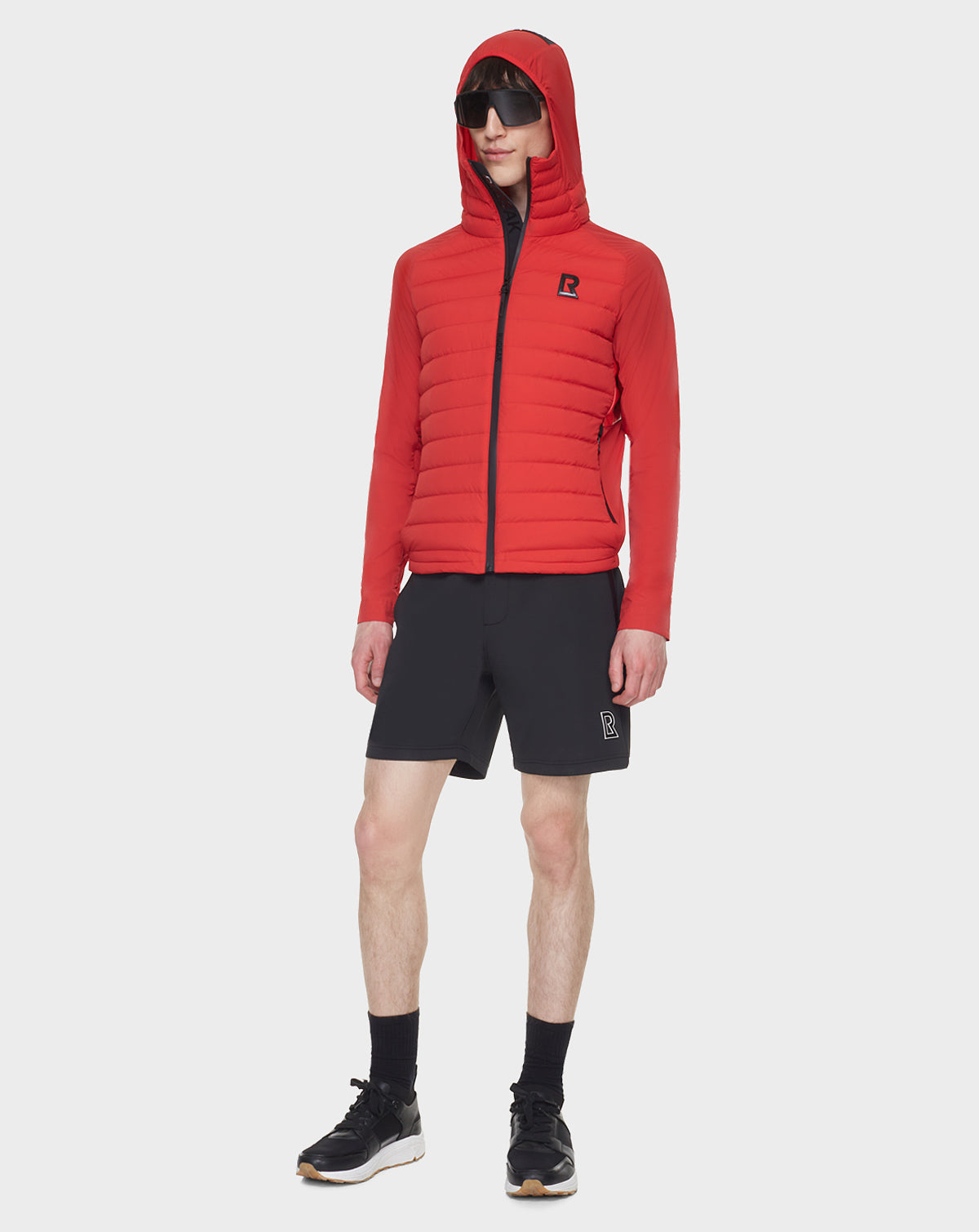 A person stands against a white background wearing RUDSAK’s JARRY - 6122007 POPPY RED water-resistant jacket, black shorts, black socks, black sneakers, and sunglasses.