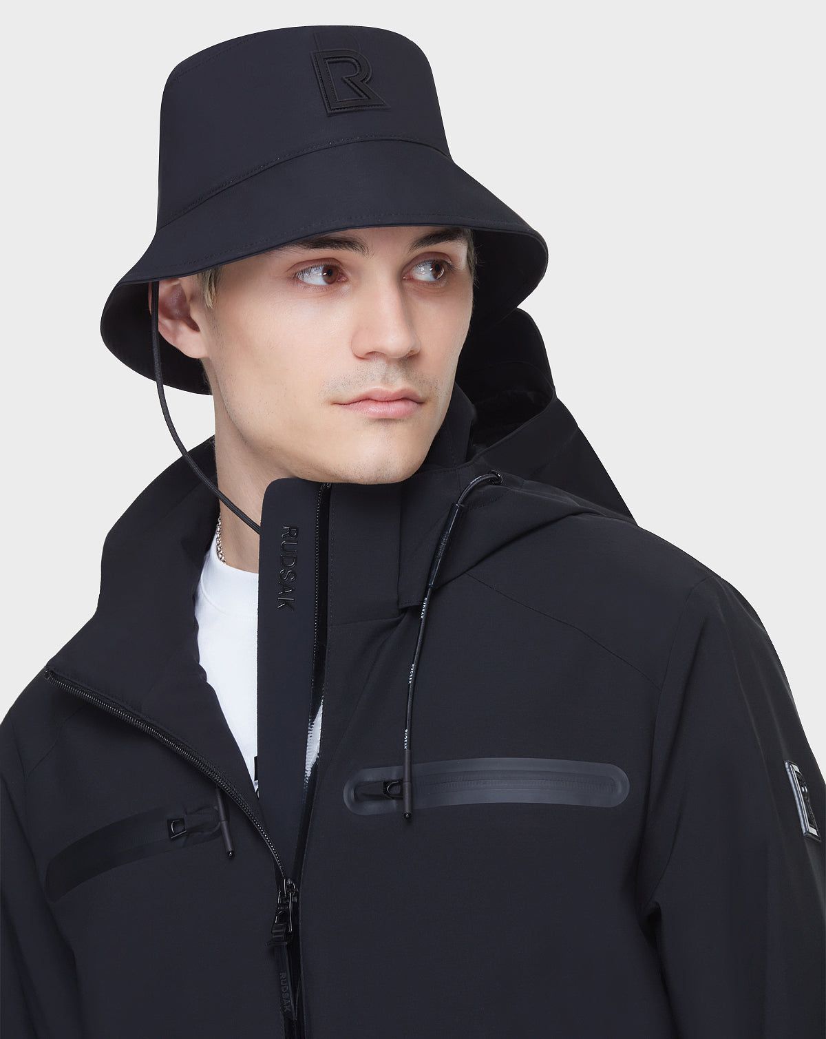 Wearing the RUDSAK JACY - 6122000 BLACK bucket hat with raised R logo and a mid-length rainwear jacket with zipper details, a person looks to the side against a plain light background, paired with a white shirt.