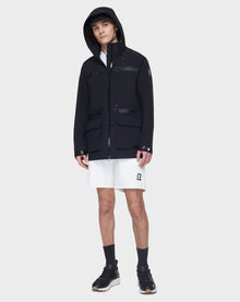A person wearing the RUDSAK JACY - 6122000 BLACK rainwear jacket, white shirt, white R-logo shorts, black socks, and black sneakers stands against a plain light gray background.