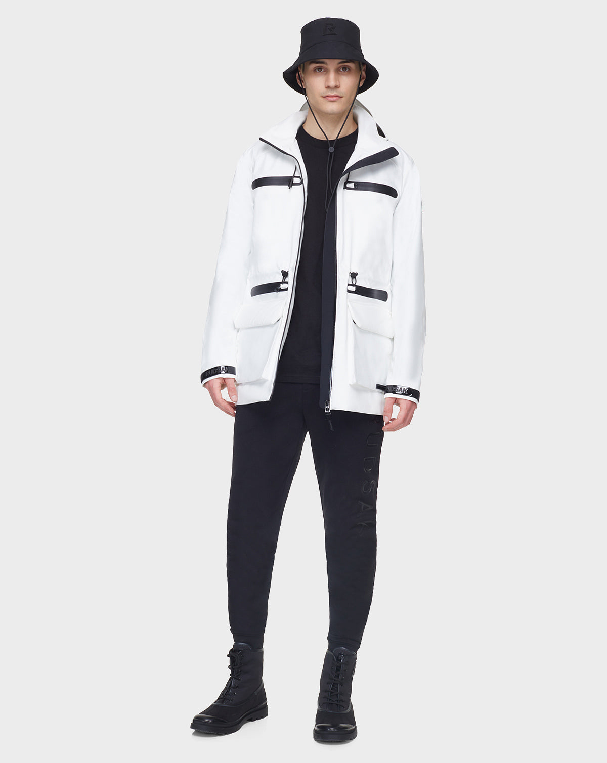 A person stands facing forward against a light background, wearing all black with the RUDSAK JACY - 6122000 WHITE raincoat, a white mid-length coat featuring multiple black zippers.